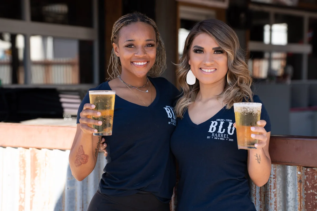 Two Blu Barrel staff members in navy uniform shirts raising cold beers on the patio